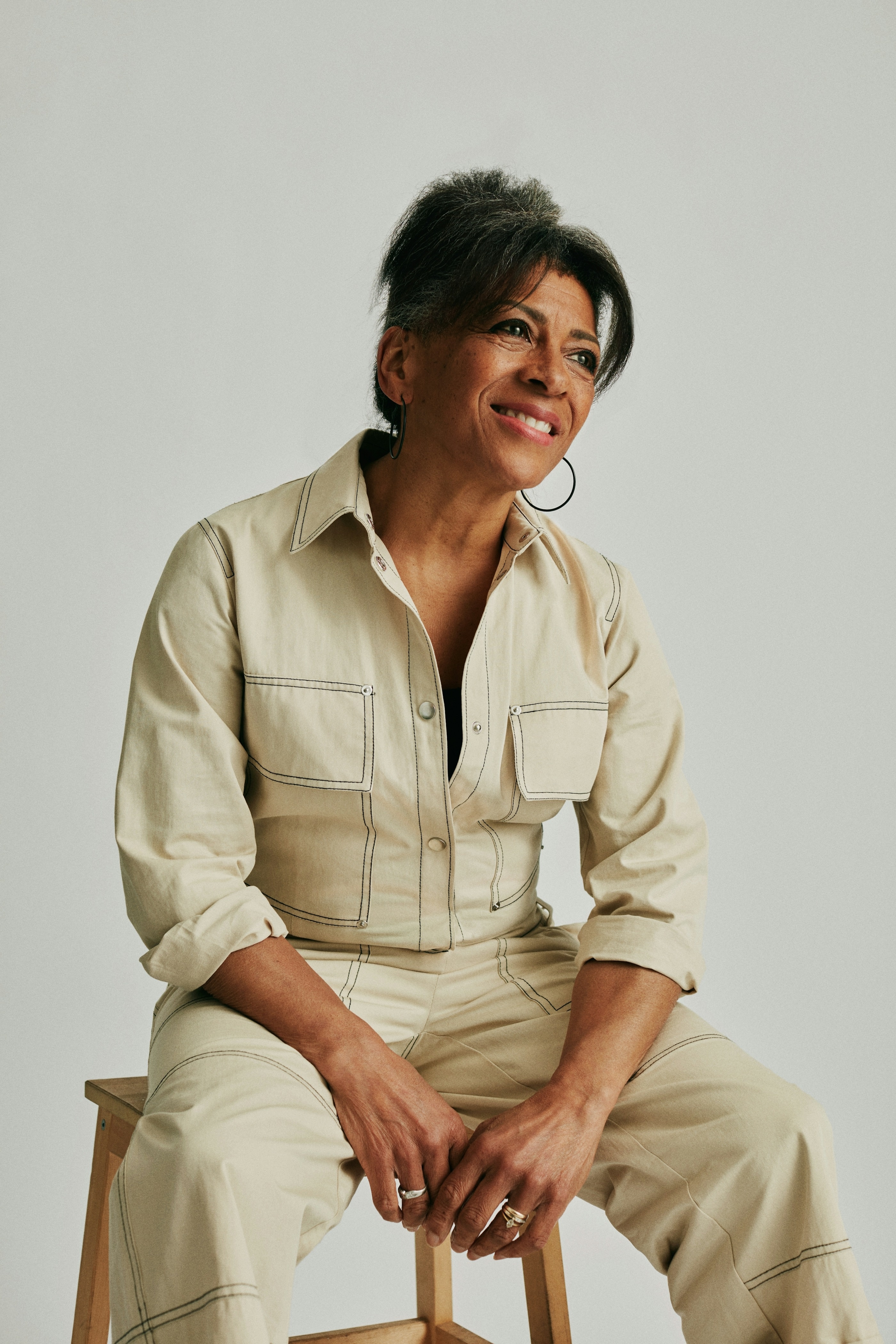 Woman in a beige jumpsuit smiling while seated on a stool